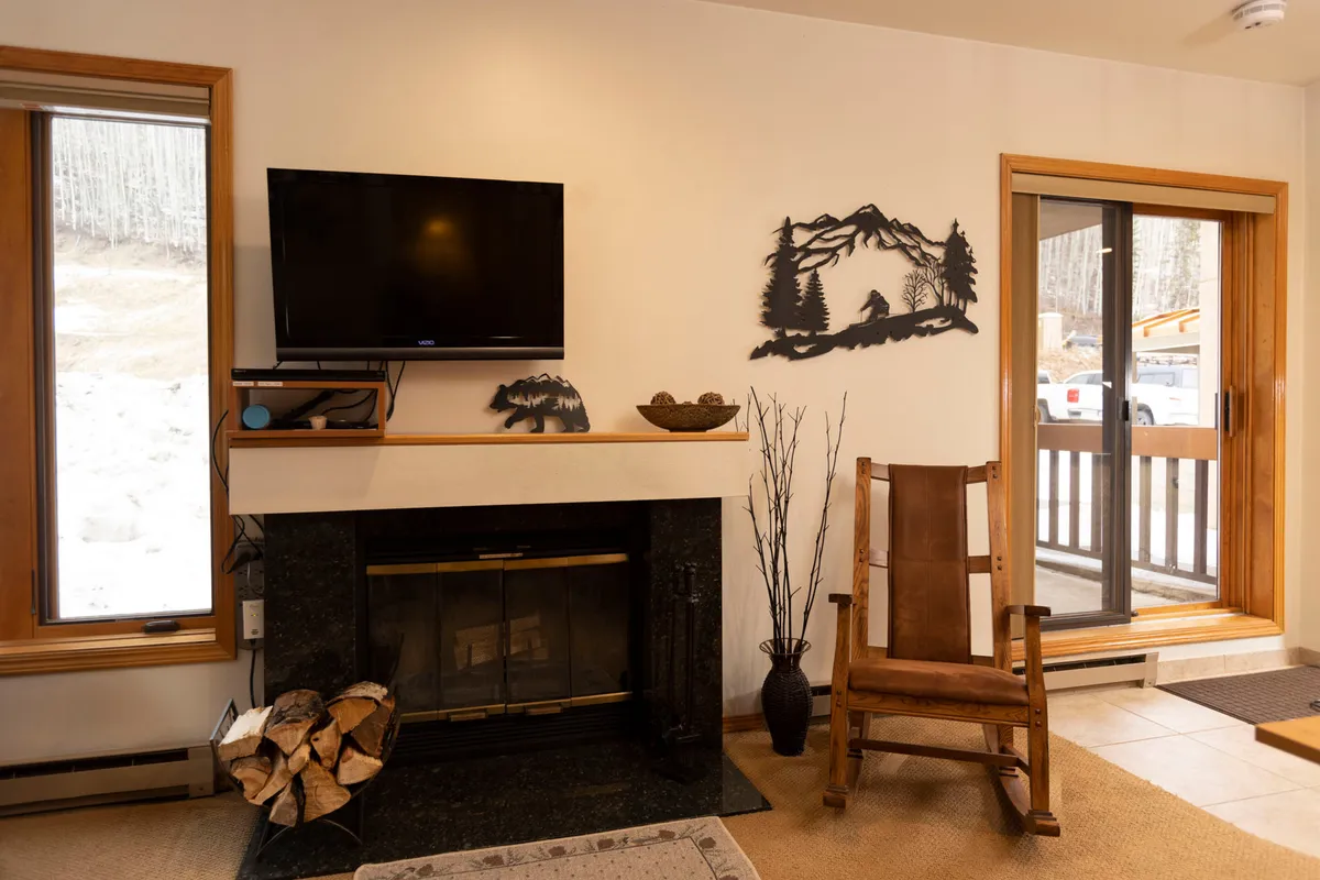 A welcoming nook centered on a glowing fireplace, with a rocking chair, a tidy stack of wood, a TV, and thoughtful wall art. The space is styled for maximum comfort, suggesting cozy evenings spent reading or sharing stories by the fire.