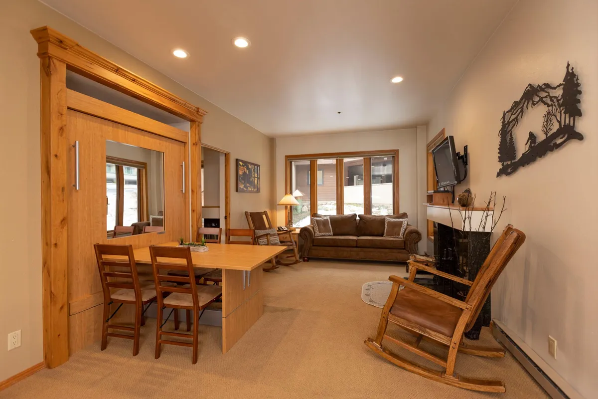 The image showcases a warm, inviting living area with a comfortable couch, a stylish dining table, a classic rocking chair, and a glowing fireplace. Large windows bathe the space in natural light, creating a bright and airy ambiance. The setting hints at cozy evenings by the fire and relaxed mornings soaking in the mountain views.