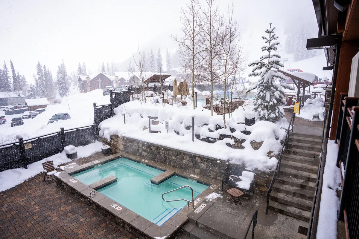 A steaming hot tub surrounded by snow, bordered by evergreen trees and rustic stone steps. The setting is serene and luxurious, with the winter landscape enhancing the spa-like experience. This is an ideal spot for stargazing or enjoying a sunset soak after a day on the slopes.
