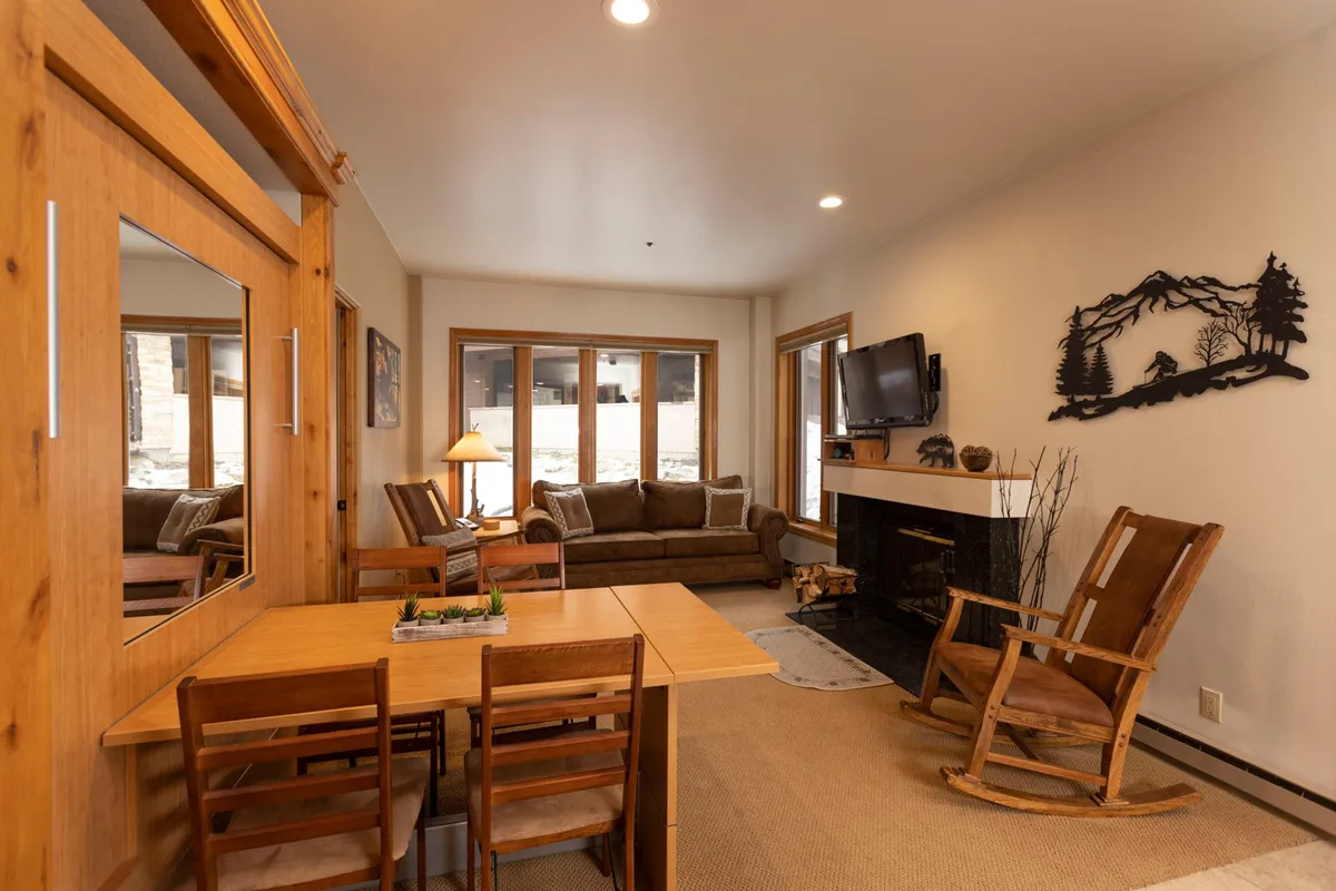 A spacious open-plan living and dining area featuring a cozy sofa, a central fireplace, a TV, a rocking chair, and a large dining table. Light floods the space from panoramic windows, and the arrangement invites socializing, relaxation, and family meals. The dining area’s proximity to the living room highlights the space’s flexibility and togetherness.