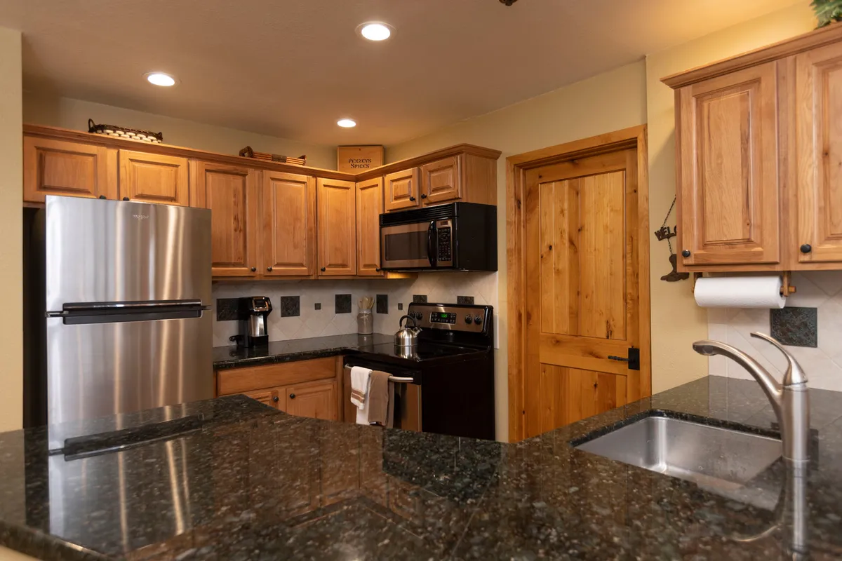 This angle focuses on the kitchen’s stainless steel refrigerator, black stove and microwave, granite countertop, and wooden cabinetry. The scene feels both cozy and functional, ideal for culinary experiments and shared meals.