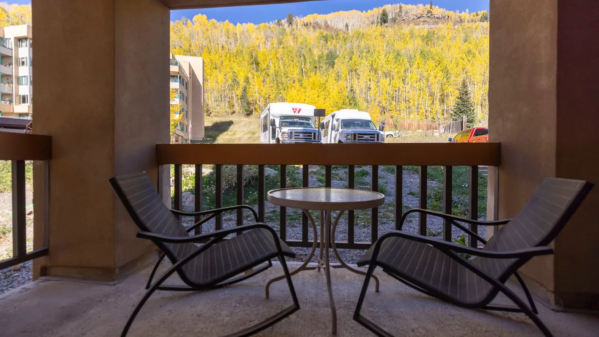 A serene exterior patio scene featuring two lounge chairs and a round table set amidst colorful autumn trees. The setup is ideal for peaceful moments, whether enjoying morning coffee, reading a book, or sipping wine at sunset. The ambiance is calm, intimate, and perfectly suited for couples looking for a romantic outdoor setting.