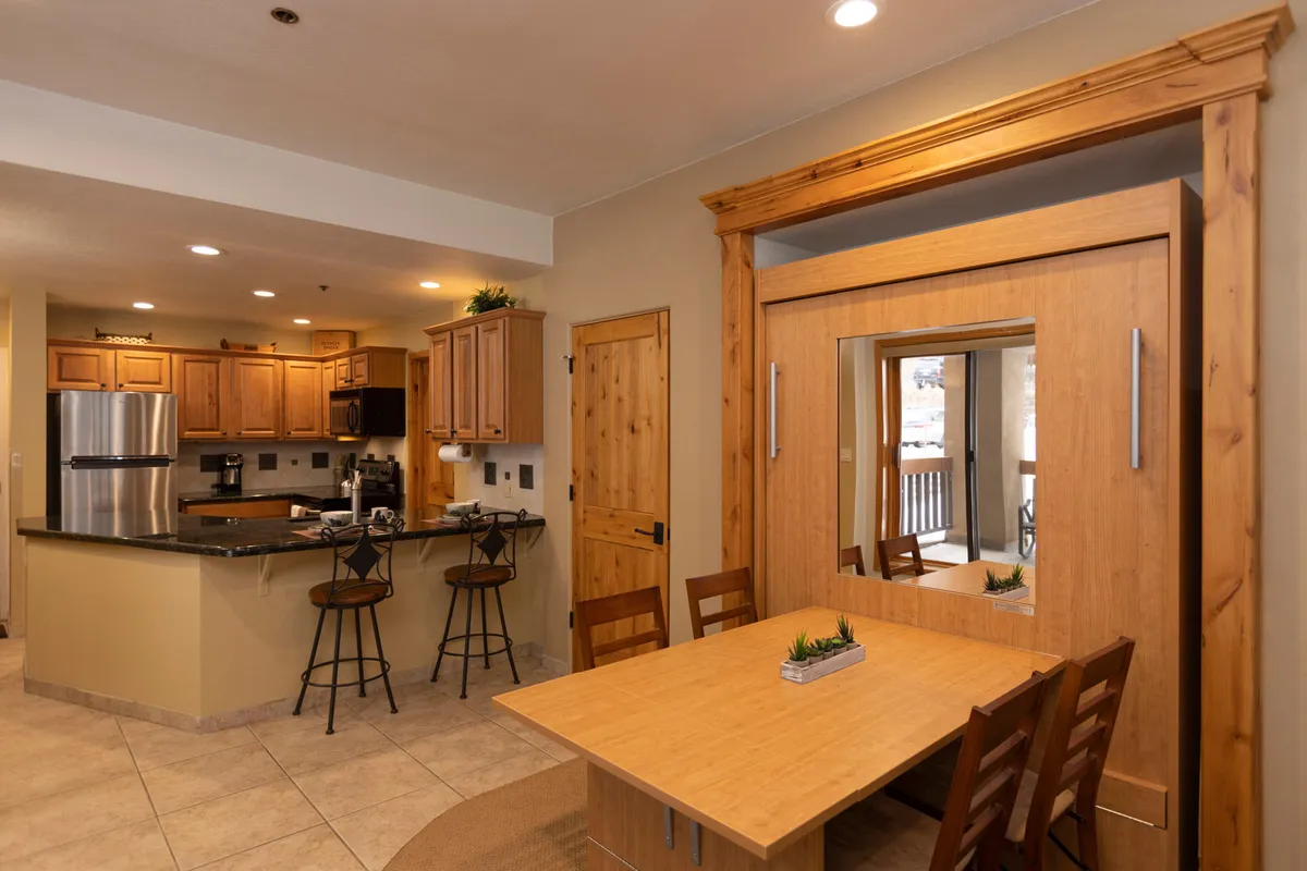 This image showcases a cozy kitchen and dining area, featuring a kitchen island, wooden cabinets, a dining table, and a mirror. The scene feels welcoming and is set up for both cooking and shared meals, with space for socializing and relaxing.