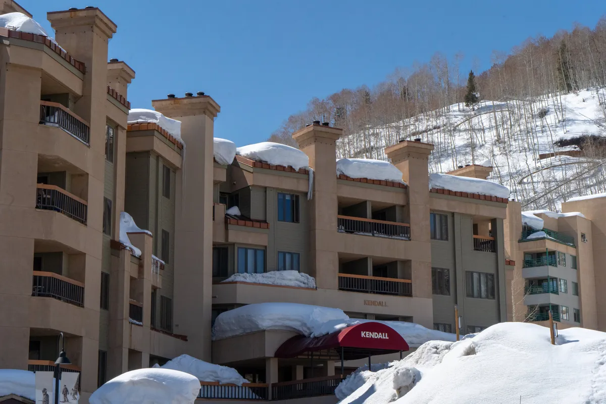 The image shows the building facade of the condo, snow-covered roofs, and a welcoming entrance awning. The scene is lively yet cozy, with sunlight glinting off the snowy surroundings, suggesting an inviting, family-friendly environment close to the slopes and village amenities.