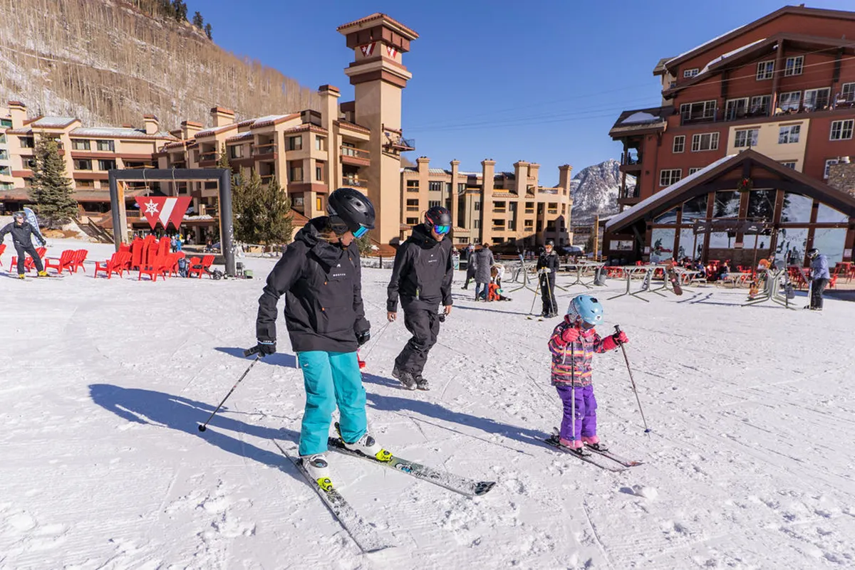 A playful winter scene with a family skiing together in front of the snow-covered resort building. The image radiates energy and togetherness, emphasizing making memories on the slopes and easy access from accommodation to mountain activities.