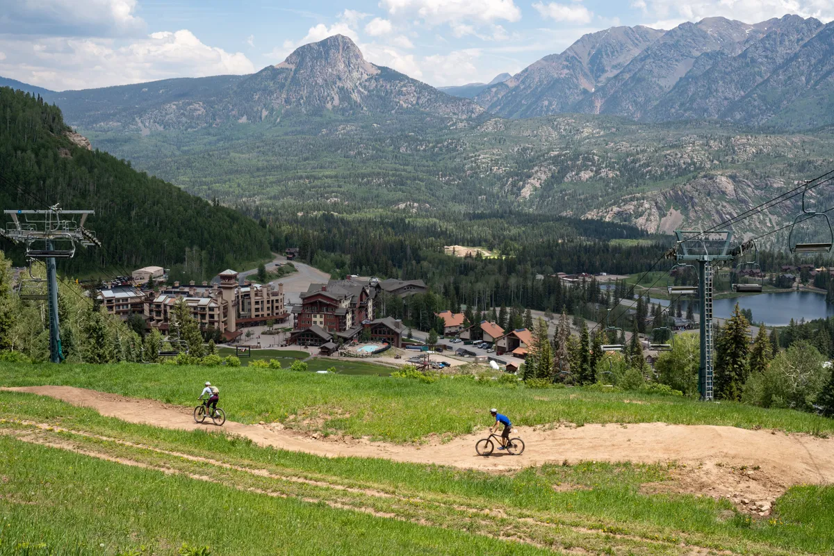 The image features a sweeping mountain range, bikers on a nearby trail, resort buildings, and a ski lift. It communicates excitement and possibility, emphasizing activities like mountain biking, hiking, skiing, and exploring nature—all key attractions for family and group vacations.