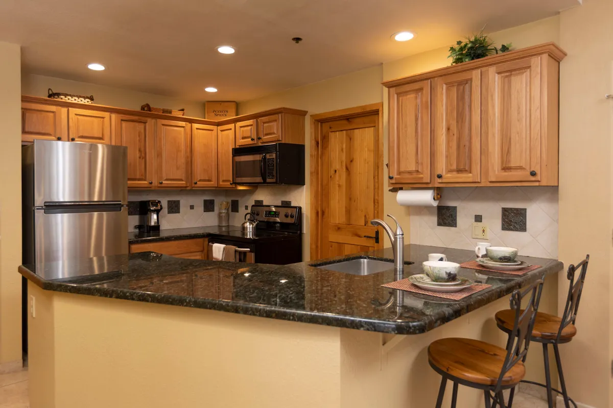 The kitchen features elegant granite countertops, wood cabinetry, stainless steel appliances, and bar stools. The space is designed for cooking, entertaining, and casual family meals, with a warm and homely mood.